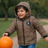 Happy toddler boy in brown hooded winter jacket playing outside with orange ball on a green path