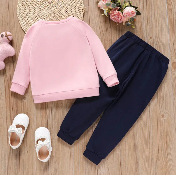 Toddler outfit with pink long-sleeve sweatshirt, navy blue pants, white shoes, and plush llama toy on wooden floor